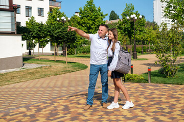 Back to school. Smiling father proudly hugs his schoolgirl daughter with backpack and uniform on the street and pointing to school with his hand.