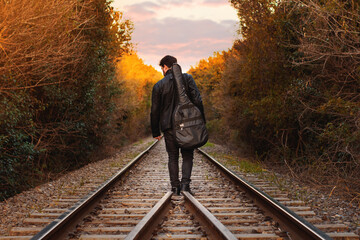 Hombre apuesto con chaqueta de cuero  y guitarra al hombro caminando por vías del tren - obrazy, fototapety, plakaty