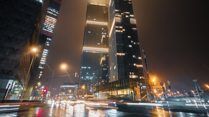 Busy intersection in Moscow city center at night. Bottom view to the cars passing by with bright light trails and overhanging skyscrapers glittering in raindrops against illuminated sky. © railwayfx