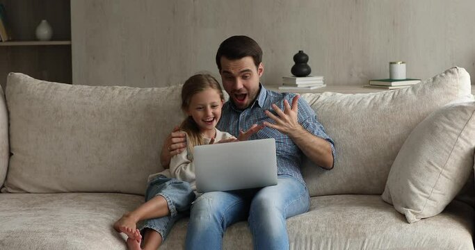Young father and little adorable 8s daughter sit on comfy couch at living room using laptop enjoy weekend free time on internet, watching together cartoons on-line, enjoy video-sharing website content