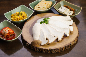 Fresh, raw sliced Eringi or King Oyster Mushroom on wooden tray with kimchi side dish and vegetable. Vegetarian, vegan and meatless. Korean BBQ or Japanese Yakiniku Barbecue concept. Selective focus.