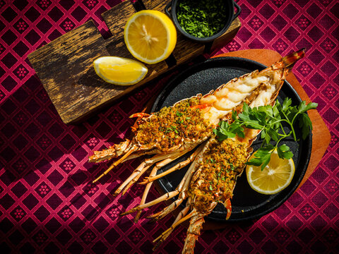Delicious Spiny Lobster halved and grilled with lemon and Italian parsley herb bread crumbs on cast iron skillet with wooden serving plate on geometric magenta background. Close Up. Modern setting.