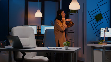 Fototapeta premium Exhausted businesswoman gesturing while talking at phone discussing marketing strategy late at night in startup office. Manager negotiating management statistics analysing company consultation ideas