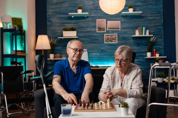 Elderly couple playing chess on board sitting at home together on couch. Old mature retired man and...