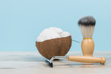 Shaving foam in a coconut bowl and wooden shaving accessories on a wooden table.