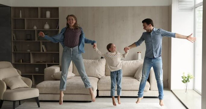 Couple and little cute daughter holding hands jumping dancing in cozy living room celebrate relocation day to new apartment. Family bonding and wellbeing at first own house, tenancy, mortgage concept
