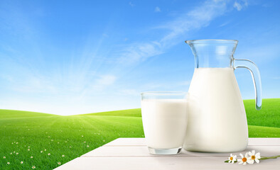 Glass and pitcher of fresh milk on white wooden table with green meadow background.