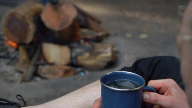 Blonde male drinking cup of tea and enjoy bonfire at forest campsite, back view