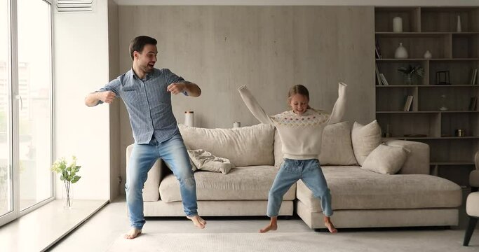 Young cheerful man have fun enjoy active dances with cute little daughter in cozy living room, full-length view. Carefree leisure time together at home, family weekend funny activities, hobby concept