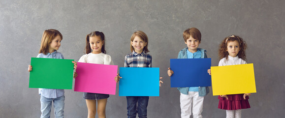 Team of 4 happy cheerful little kids showing blank mockup sign boards or posters. Group of four cute smiling children holding colorful paper banners standing against grey header studio background