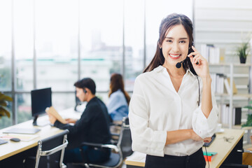 Smiling Asian woman operator wearing headset on foreground smiling and looking at the camera with blurred customer service colleagues working on background in call center.
