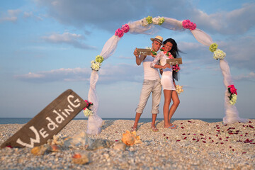 Newlywed couple in Hawaiian Hula. Groom with bride wearing lei, standing under archway on beach