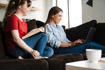 Girl with down syndrome reading book while sitting on sofa with her mother