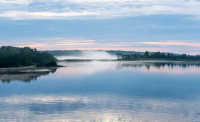 Beautiful landscape on the river covered with morning haze with reflection of clouds in the water.