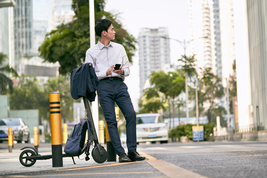 young asian business man commuter standing on downtown street with a electric push scooter
