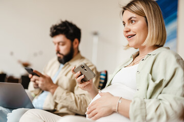 Fototapeta premium Bearded man and pregnant woman using cellphones while sitting on couch