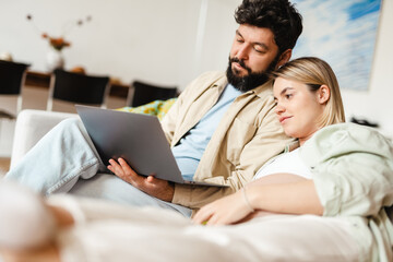 Bearded man and pregnant woman using laptop while sitting on couch
