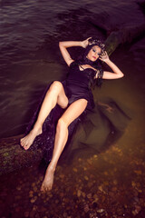 Sensual Beautiful Brunette Female In Black Dress Posing At Sea On Tree While Wearing Seashell Decorated Crown And Long Black Pearl Necklace.