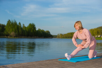 Outdoor Sport Concepts. Mature Caucasian Woman During Yoga Stretching Training On Wooden Stage Near Water Outdoor.