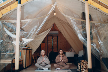 Obraz na płótnie Canvas Woman senior and young relaxing at glamping camping tent. Women family elderly grandmother and young granddaughter doing yoga and meditation indoor. Modern zen-like vacation lifestyle concept