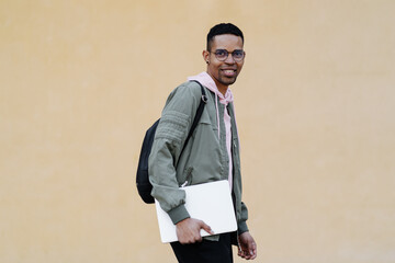 African man in glasses holding laptop on pale yellow background. Shot of carefree black male freelancer smiling to camera.