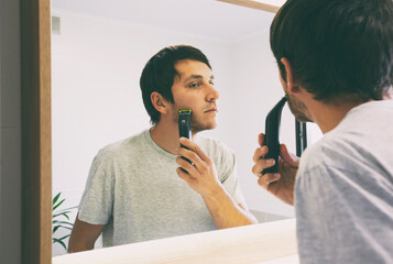 Young guy shaving with electric razor in the bathroom in front of the mirror