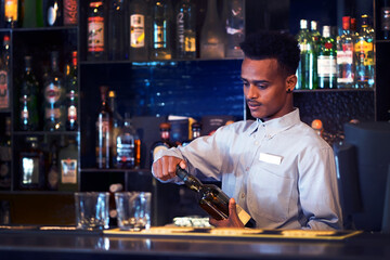 African young bartender opens a bottle of whiskey. Shelves with bottles of alcohol in the background.