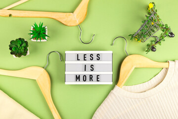 Less is more - written on lightbox next to Shopping cart entwined with plants on green background among wooden hangers. Conscious consumption slow fashion Zero waste concept. Top view flat lay - obrazy, fototapety, plakaty
