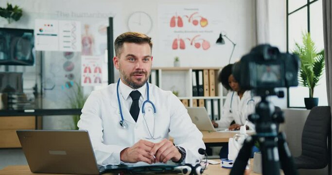Attractive confident experienced bearded doctor sitting in front of camera in own medical office and record video for his internet audience