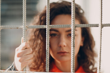 Young brunette curly woman in orange suit behind jail bars. Female in colorful overalls portrait - obrazy, fototapety, plakaty