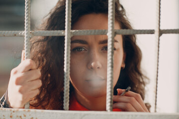 Young brunette curly woman in orange suit behind jail bars smokes cigarette. Female in colorful...