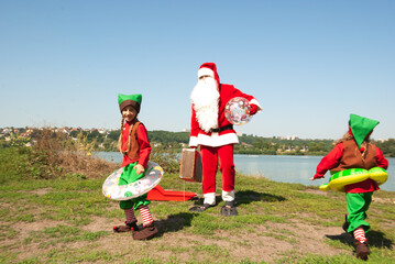 Santa Claus with a suitcase and elf children in rubber rings dance on the beach by the water for Christmas. winter hotel vacation concept. Advertising for the new year