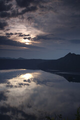 山間の湖の湖面に映る太陽の輝く曇り空。