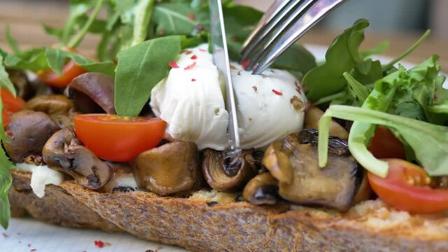 Fork and knife cut poached egg and yolk flows out on canape with mushrooms tomatoes and ruccola on piece of wholemeal bread macro