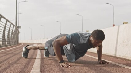 Slowmo shot of athletic African-American man in sportswear and wireless headphones doing clapping push-ups training outdoors on race track on summer morning