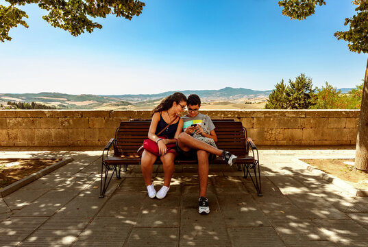 Girl and Boy smiling and looking at the mobile phone sitting on a park bench in belvedere of San Casciano dei Bagni, Siena