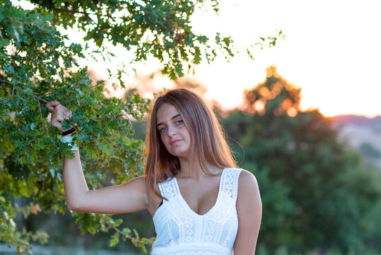 A young girl with blond long hair, dressed in white, poses at sunset touching the leaves of the magic tree
