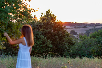 Blurred sunset profile of a young girl with long blonde hair dressed in white as she touches the...