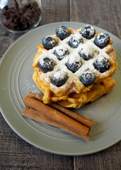 Belgian gluten-free pumpkin waffle, sprinkled with powdered sugar and blueberries, lies on a gray plate with cinnamon sticks on a wooden table, side view. vegan breakfast prepared at home