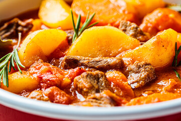 Beef stew with potatoes and carrots in tomato sauce in red pot, gray  background.