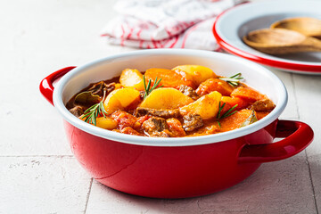 Beef stew with potatoes and carrots in tomato sauce in red pot, gray  background.