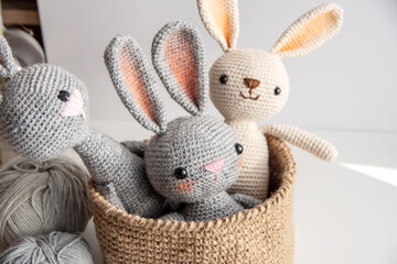 Three handmade bunnies sit in a jute basket on a white table, knitting yarn side by side.