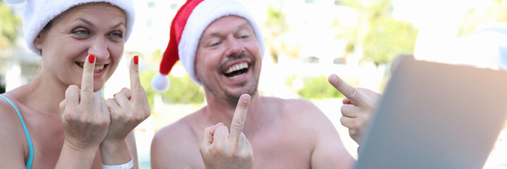 Joyful man and woman in red santa claus hats show gesture of fact in laptop monitor being at resort...