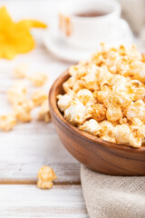 Popcorn with caramel in wooden bowl and a cup of coffee on a white wooden background. Side view, selective focus.