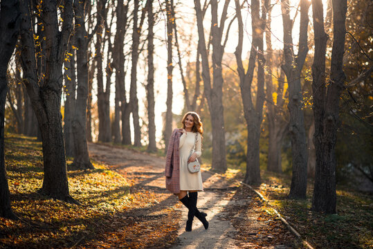 beautiful young blonde woman walking through the autumn city in a stylish outfit. Knitted white woolen dress, high black boots. Fashionable plaid coat.