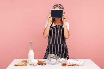 Image of young lady in striped apron and covering her face with tablet computer with black blank screen, cooking homemade pastry, ingredients. Indoor studio shot isolated on pink background.