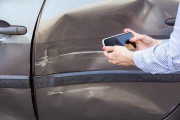 Unknown woman with mobile phone in hands taking picture of damaged part of car, photographing...