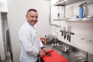 Naklejka na ściany i meble Cheerful male chef smiling to the camera while working at his kitchen