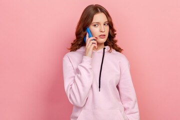 Portrait of serious curly haired teenage girl in hoodie using mobile phone, making call and talking with friend, connection. Indoor studio shot isolated on pink background