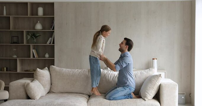 Young father holds hands little daughter while she jumping on soft comfort sofa in cozy living room. Active cheerful family spend free time together having fun enjoy playtime at home. Leisure concept